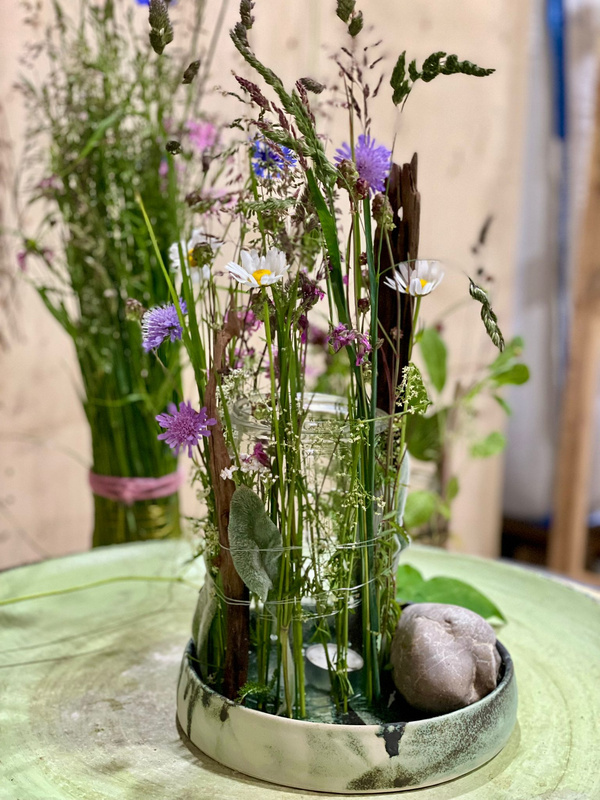 KI generiert: Ein Arrangement aus Wildblumen und Gräsern in einer Glasvase mit einem Stein auf einem grünen Tisch.