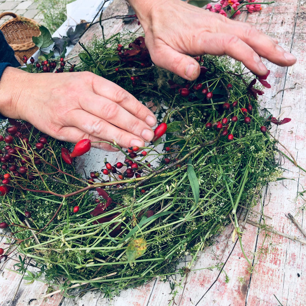 KI generiert: Hände gestalten einen Kranz aus grünen Zweigen und roten Beeren auf einem Holztisch.