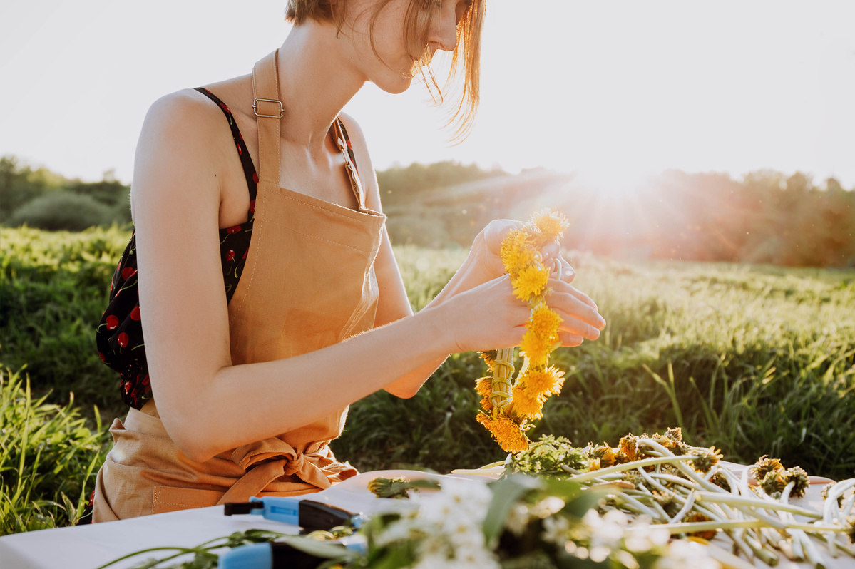 KI generiert: Eine Person bindet einen Blumenkranz aus gelben Blumen auf einem Tisch im Freien bei Sonnenuntergang.