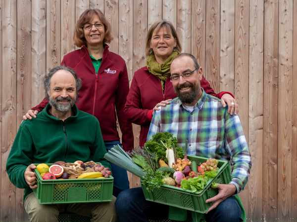 KI generiert: Vier Personen posieren mit Kisten voller frischem Obst und Gemüse.