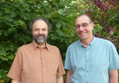 KI generiert: Zwei lächelnde Männer stehen vor einem grünen, bewachsenen Hintergrund.