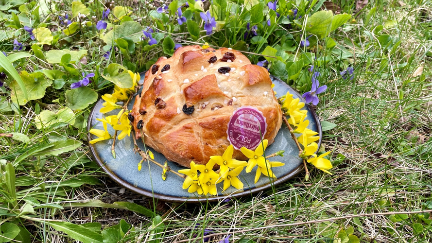 KI generiert: Ein Brot mit Rosinen auf einem Teller, umgeben von Blumen im Gras. Text: "Osterfladen 7,00 €".