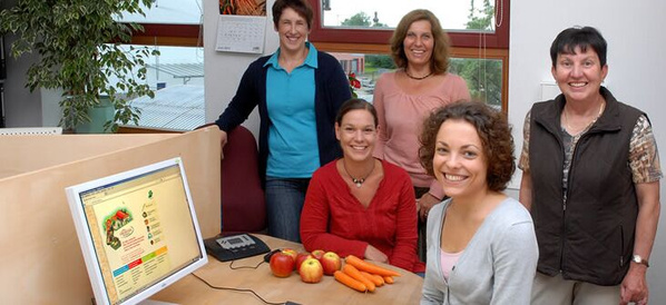 KI generiert: Fünf Personen posieren in einem Büro mit Obst und Gemüse auf einem Tisch. Ein Computerbildschirm zeigt eine Webseite.