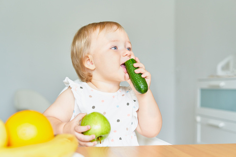 KI generiert: Ein Kind isst eine Gurke und hält einen Apfel, mit Bananen und Orangen im Vordergrund.