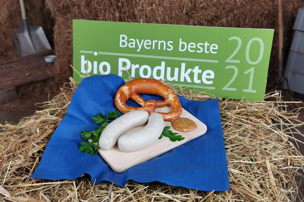KI generiert: Eine Brotzeit aus Weißwürsten und Brezel auf Stroh. Text: "Bayerns beste Bio-Produkte 2021".