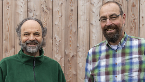 KI generiert: Zwei Männer stehen vor einer Holzverkleidung und lächeln in die Kamera.