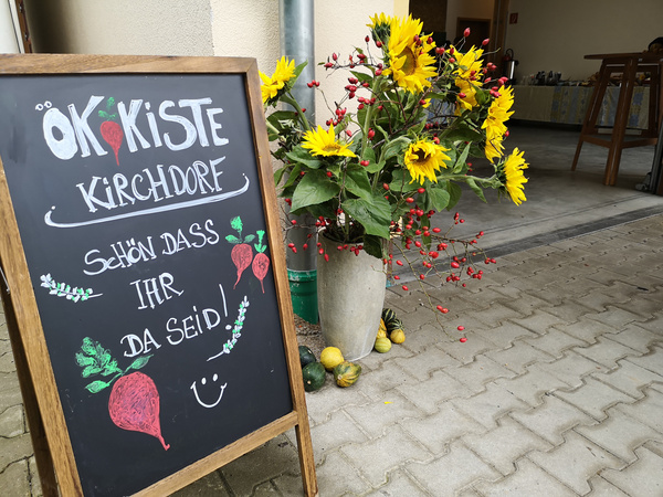 KI generiert: Tafel vor Blumenstrauß mit Text: "Ökokiste Kirchdorf - Schön dass ihr da seid!"