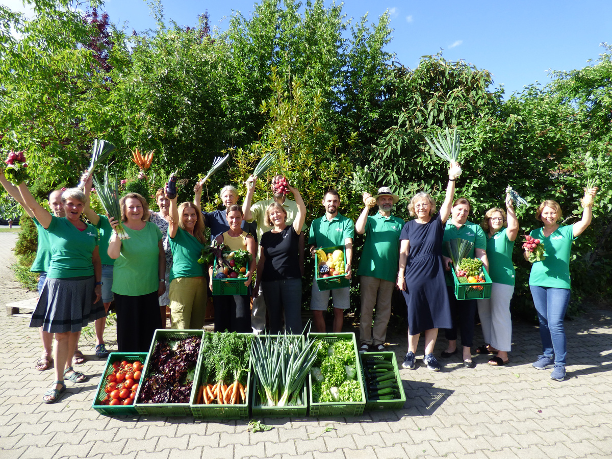 KI-generierte Beschreibung: Gruppe von Menschen mit Gemüsekisten und Blumen in grüner Kleidung.