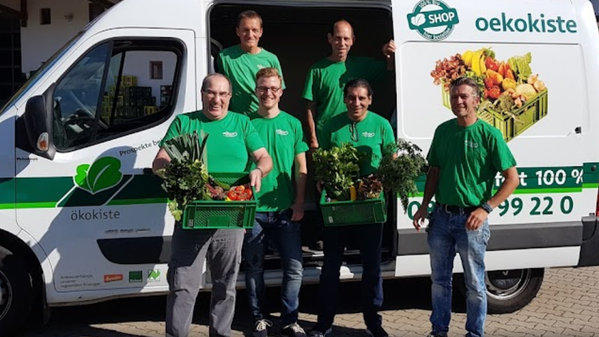 KI generiert: Mehrere Personen in grünen Shirts vor einem Lieferwagen mit Kisten voller Gemüse. Text: „oekokiste“.