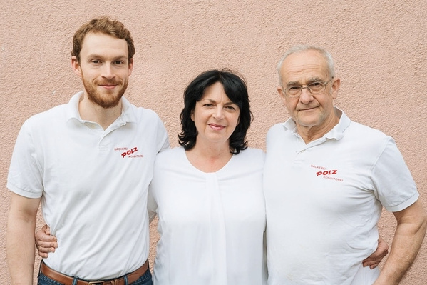 KI generiert: Drei Personen stehen lächelnd vor einer Wand. Text auf Hemden: "Bäckerei Polz Konditorei".