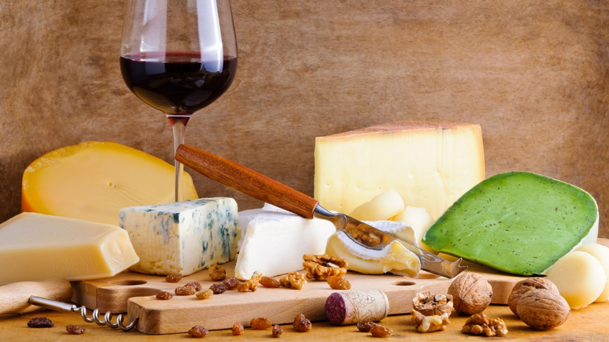 KI generiert: Verschiedene Käsesorten und ein Glas Rotwein auf einem Tisch, umgeben von Nüssen und getrockneten Früchten.