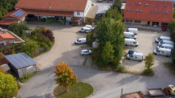 KI generiert: Luftbild eines Hofs mit Fahrzeugen, Gebäuden und Bäumen. Fahrzeuge mit Logos geparkt.