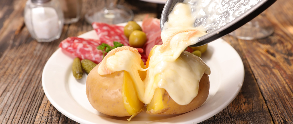 KI generiert: Geschmolzener Käse wird über eine Kartoffel gegossen, begleitet von Salami, Oliven und Gurken auf einem Teller.