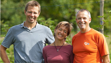 KI generiert: Drei fröhliche Personen stehen draußen, lächeln in die Kamera; im Hintergrund sind Pflanzen sichtbar.